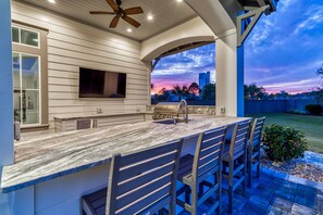 An outdoor kitchen with a marble countertop, bar stools, and a TV mounted on the wall.