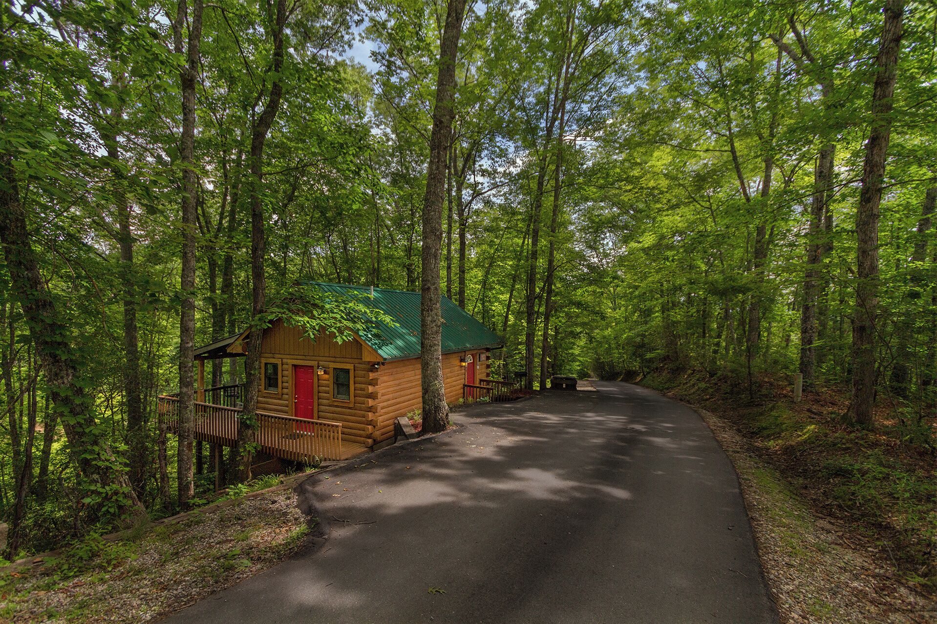 A wooden cabin with a green roof, surrounded by a forested area and a paved road.