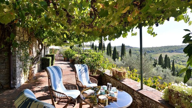 A covered outdoor seating area with wicker chairs and a glass table, overlooking a vineyard and distant hills.