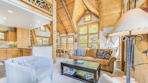 A spacious living room with a high ceiling, wooden beams, a fireplace, and a large window.