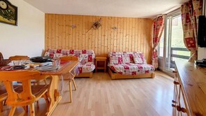 Ein gemütliches Zimmer mit Holzwänden, einem Holzesstisch mit Stühlen, zwei Betten mit gemusterten Bettbezügen und einem Fenster mit Vorhängen.