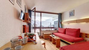 A hotel room with a wooden table, red sofa, and a large window offering a mountain view. - 100m from ski slopes, 5th floor, view ski slopes, balcony, tv, ski locker, 24m², Les Menuires (Les Menuires)