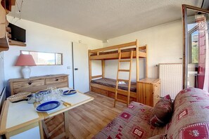 Habitación compacta con literas, una mesita con utensilios para comer, un sofá y un televisor montado en la pared.