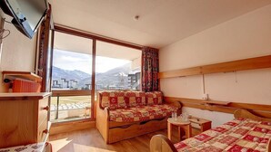 A room with a wooden bed, a sofa, a small table, and a large window offering a mountain view. - 100m from ski slopes, 2nd floor, balcony, tv, ski locker, 24m², Les Menuires (Les Menuires)