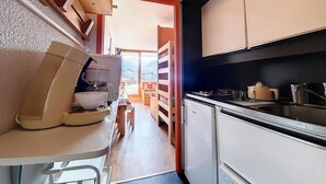 A compact kitchen with a sink, stove, and a small refrigerator. There is a wooden table with a bowl and a coffee maker. A doorway leads to a balcony with a view of buildings and mountains. - 100m from ski slopes, 2nd floor, balcony, tv, ski locker, 24m², Les Menuires (Les Menuires)