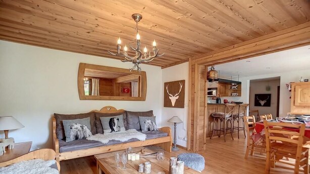 Un salon chaleureux avec un plafond en bois, un canapé agrémenté de coussins, une table basse avec des verres, et un coin repas avec des chaises et une table.