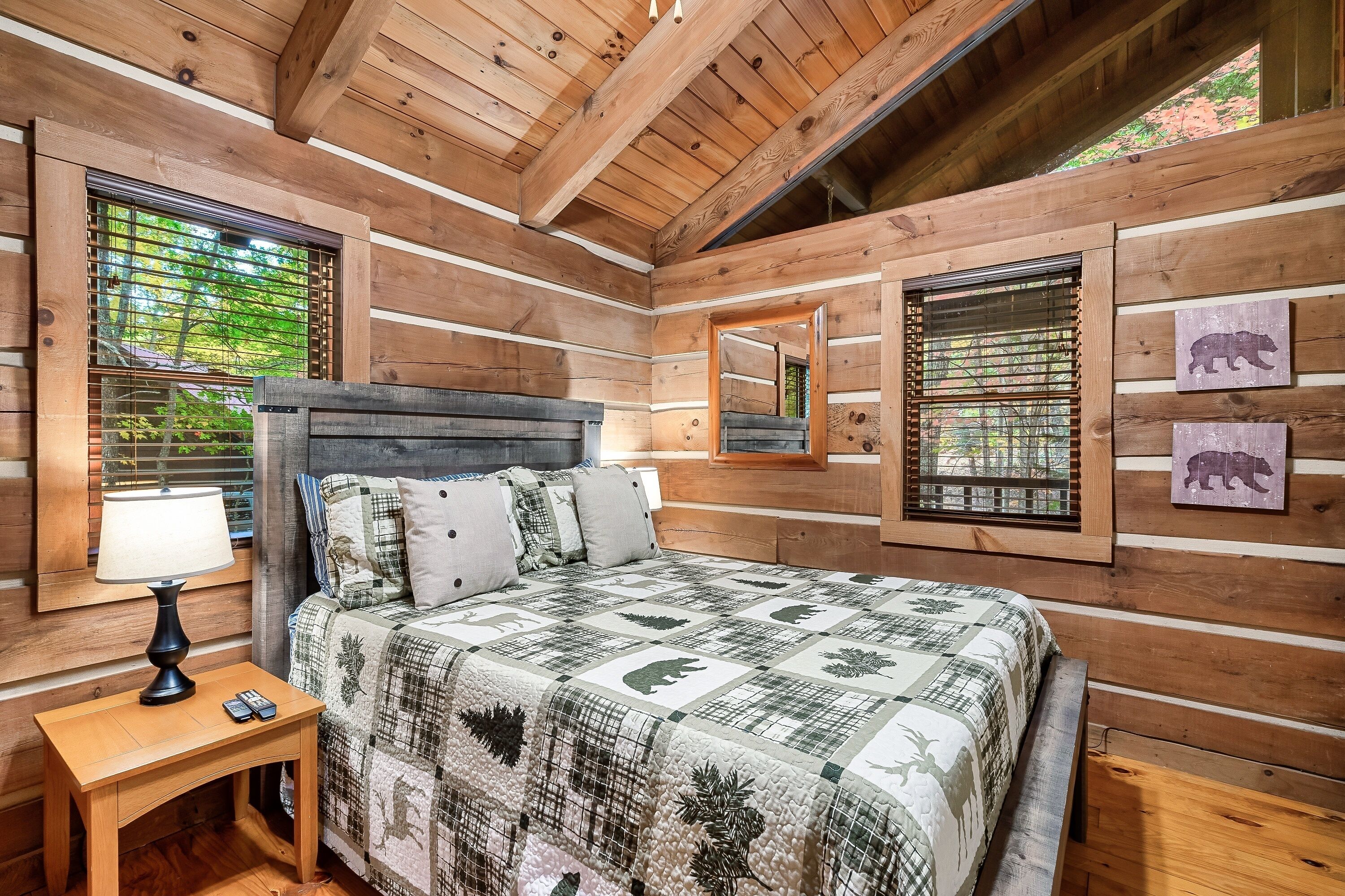 A wooden cabin bedroom with a bed, nightstand, lamp, and two windows with blinds.