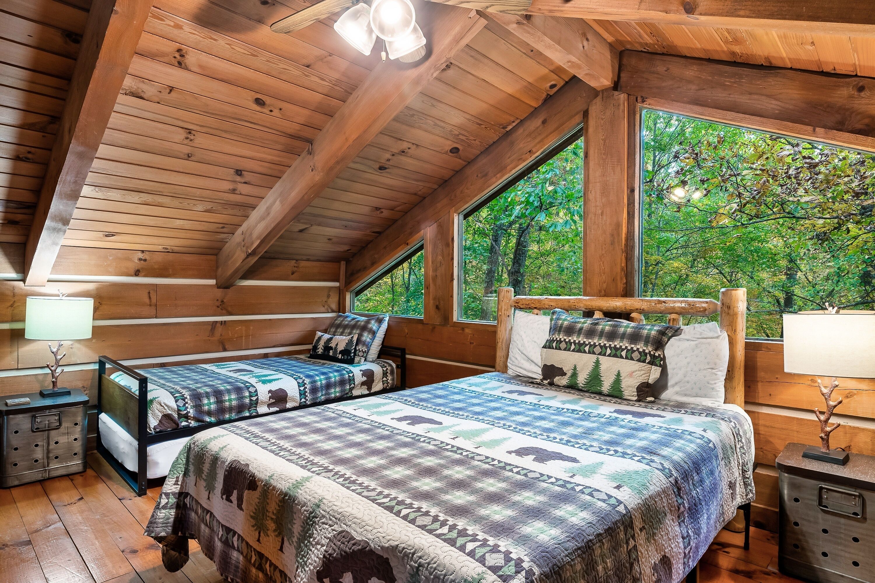 A cabin with a wooden interior, two beds with matching bedding, a nightstand, and a window with a view of trees.
