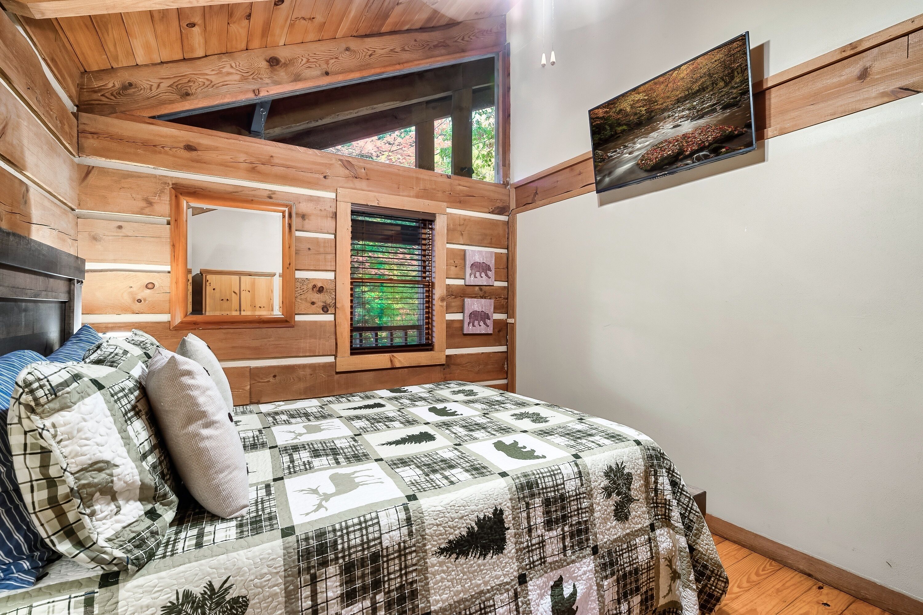 Un dormitorio con techo de madera, una cama con colcha, un espejo, una ventana con persianas y un cuadro en la pared.
