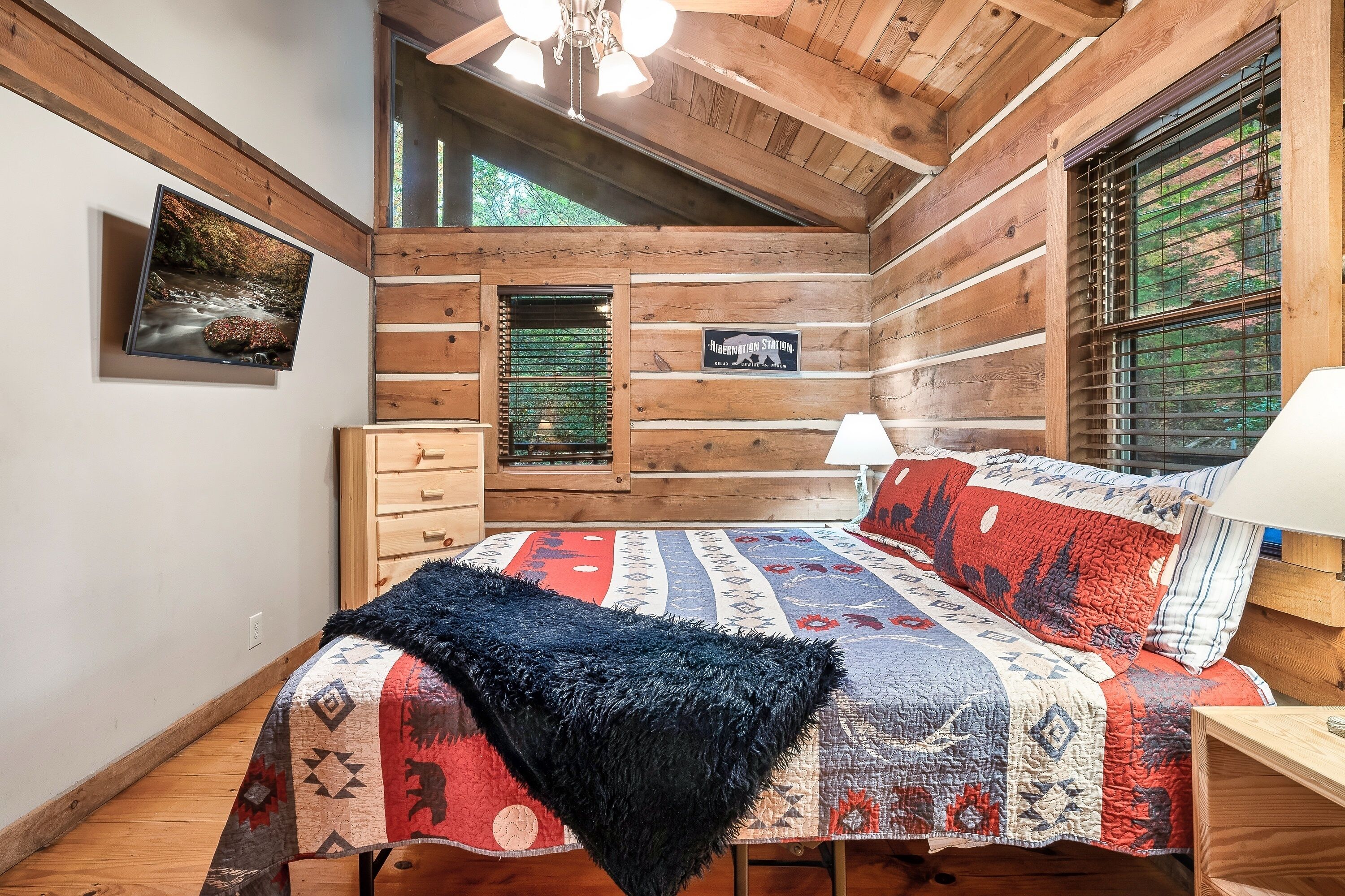 Une chambre dans une cabane en rondins, avec un lit, une commode, une fenêtre avec des stores et un ventilateur de plafond.