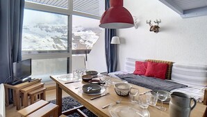 A cozy room with a wooden table set for dining, a bed with red and gray bedding, a mounted deer head on the wall, and a view of snowy mountains through the window. - 200m from resort center, 12nd floor, balcony, tv, ski locker, 37m², Les Menuires (Les Menuires)