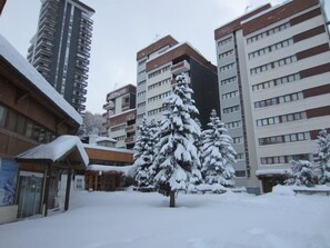 A snowy urban area with multi-story buildings, a covered bus stop, and snow-covered trees. - 50m from ski slopes, Ground floor, balcony, tv, ski locker, 19m², Les Menuires (Les Menuires)