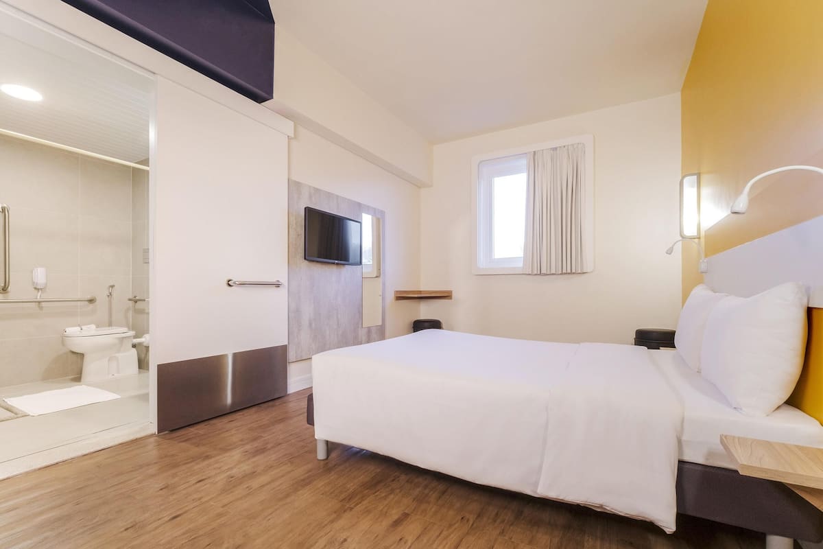 Ein Hotelzimmer mit Bett, Fernseher, Fenster mit Vorhängen, Nachttisch und Bad mit Toilette und Waschbecken.