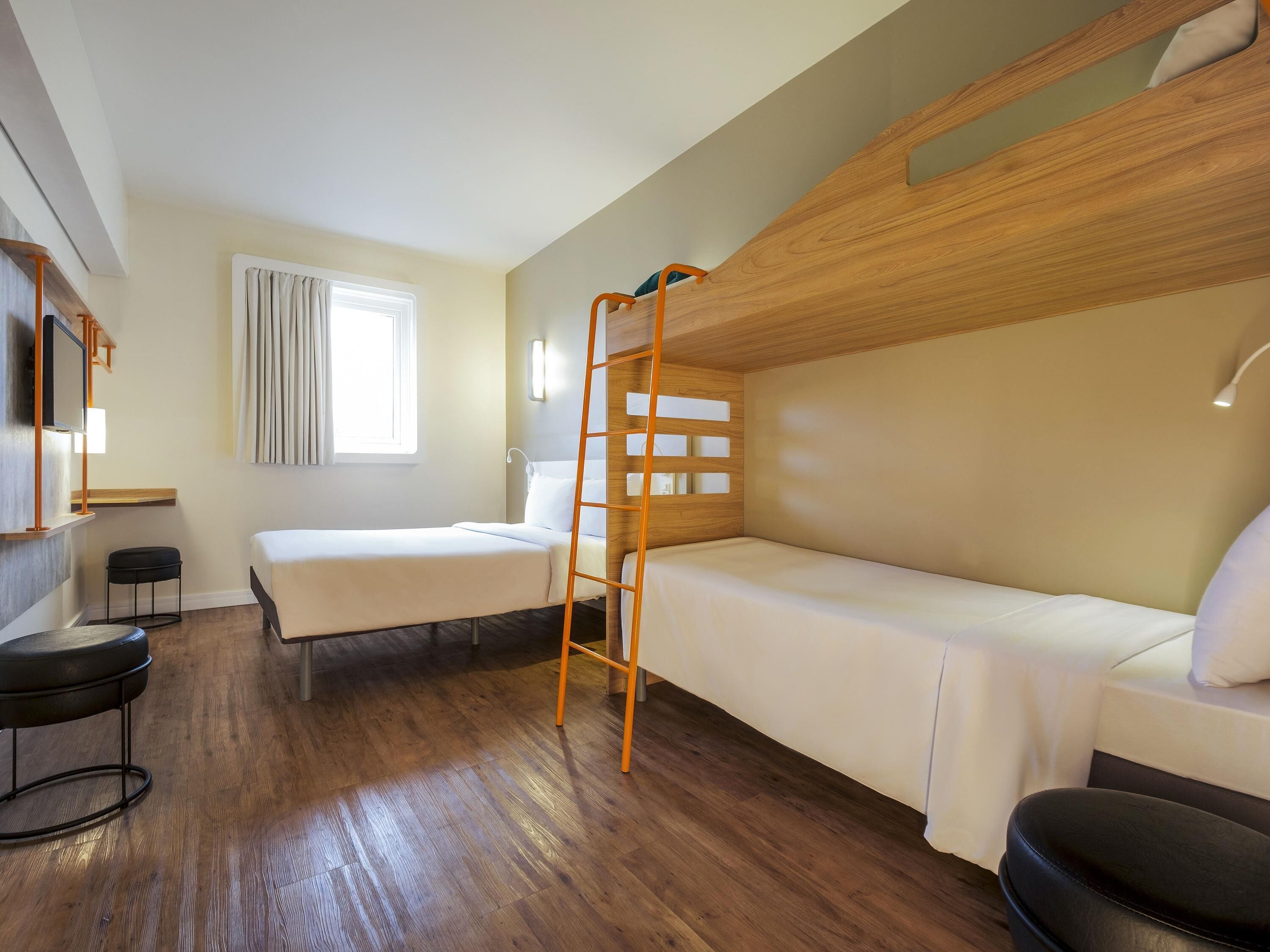 A hotel room with a bunk bed, a desk with a chair, a window with curtains, and a wooden floor.