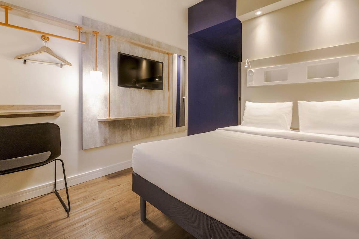 Ein modernes Hotelzimmer mit einem großen Bett, einem an der Wand montierten Fernseher, einem Schreibtisch mit Stuhl und einem Kleiderschrank.