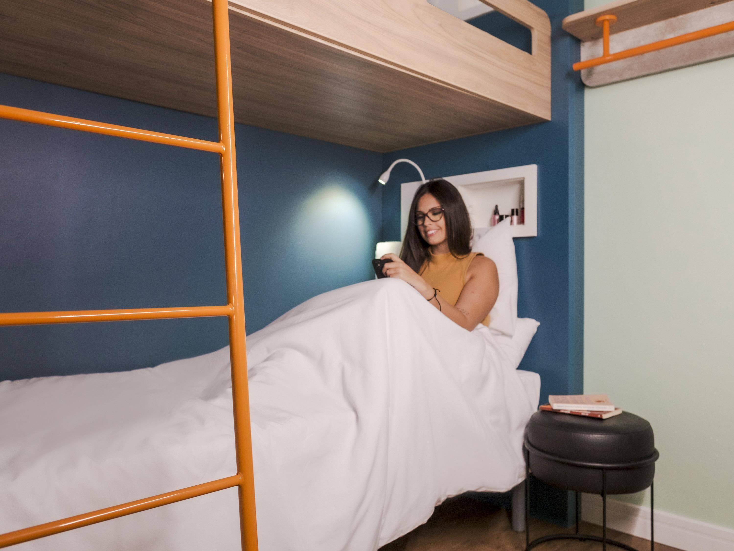 Una cama litera con escalera de madera, una mesita de noche con un libro y un estante de pared con objetos personales.