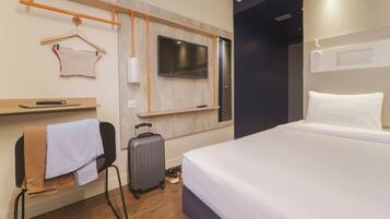 A hotel room with a bed, a TV mounted on the wall, a suitcase, a chair with a towel, and a wooden shelf.