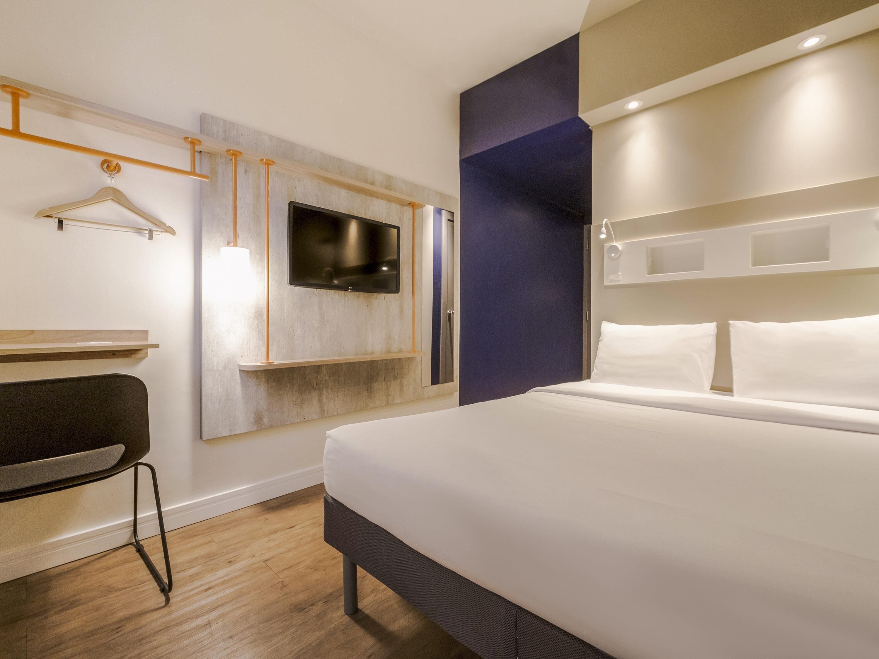Habitación de hotel con cama, escritorio, silla, televisor montado en la pared y zona de ducha.