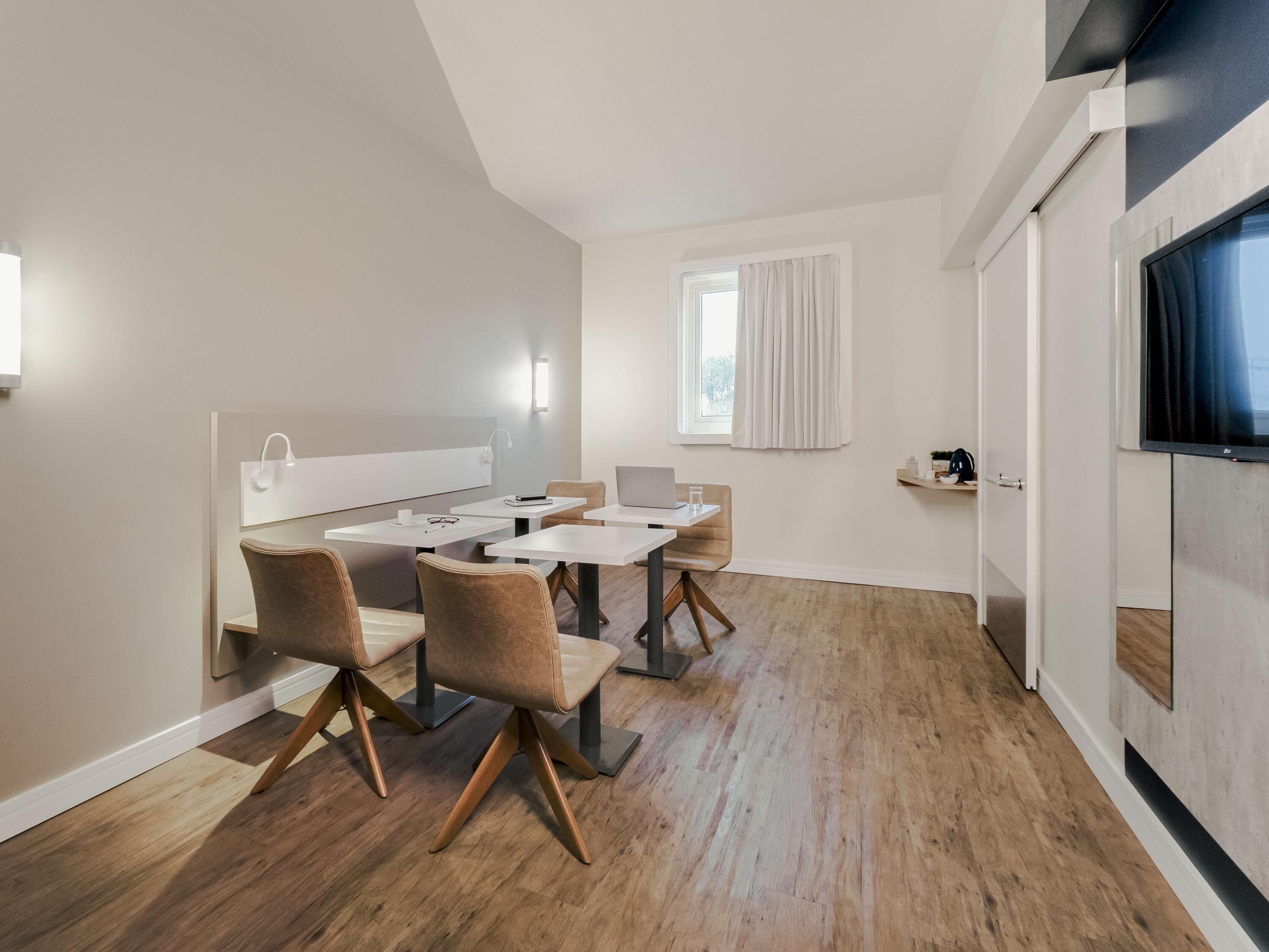 A modern, well-lit room with a dining area, a window with curtains, and a TV mounted on the wall.
