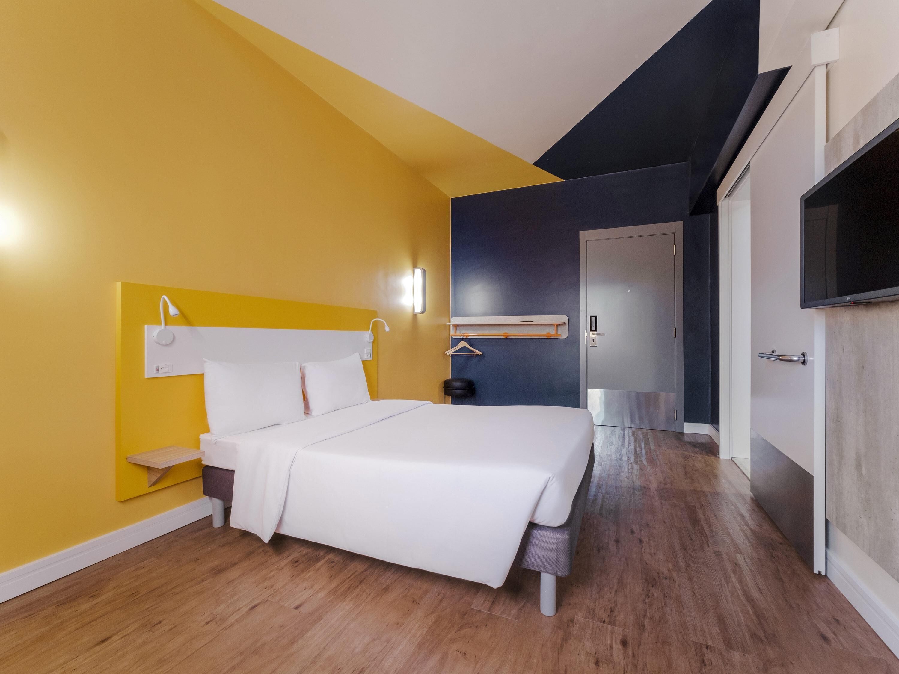 Quarto de hotel moderno com cama, mesa de cabeceira e televisão.