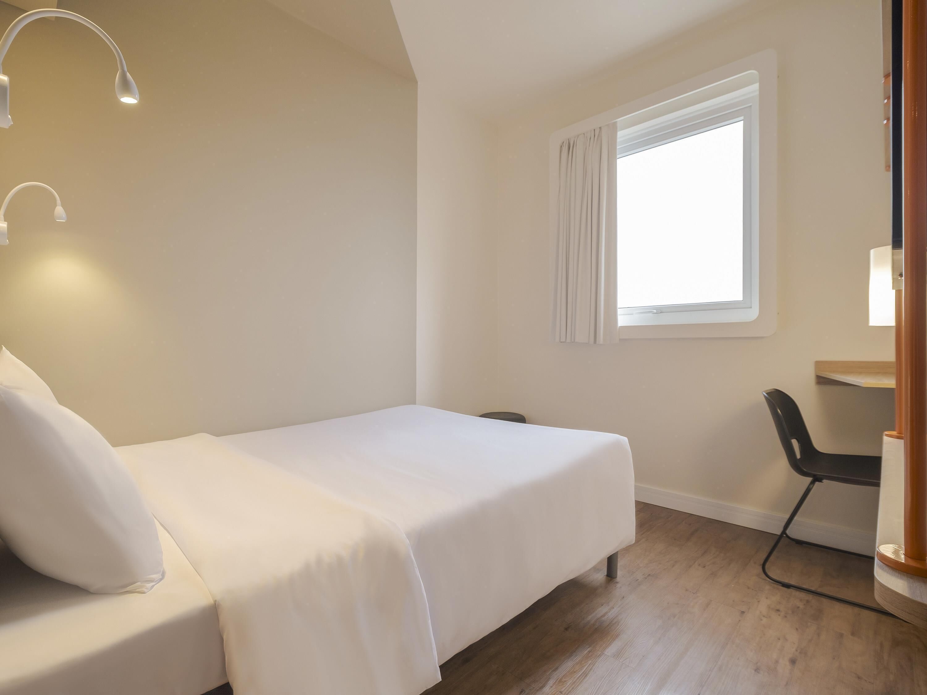 Habitación de hotel con cama, escritorio, silla, ventana con cortinas y lámpara.