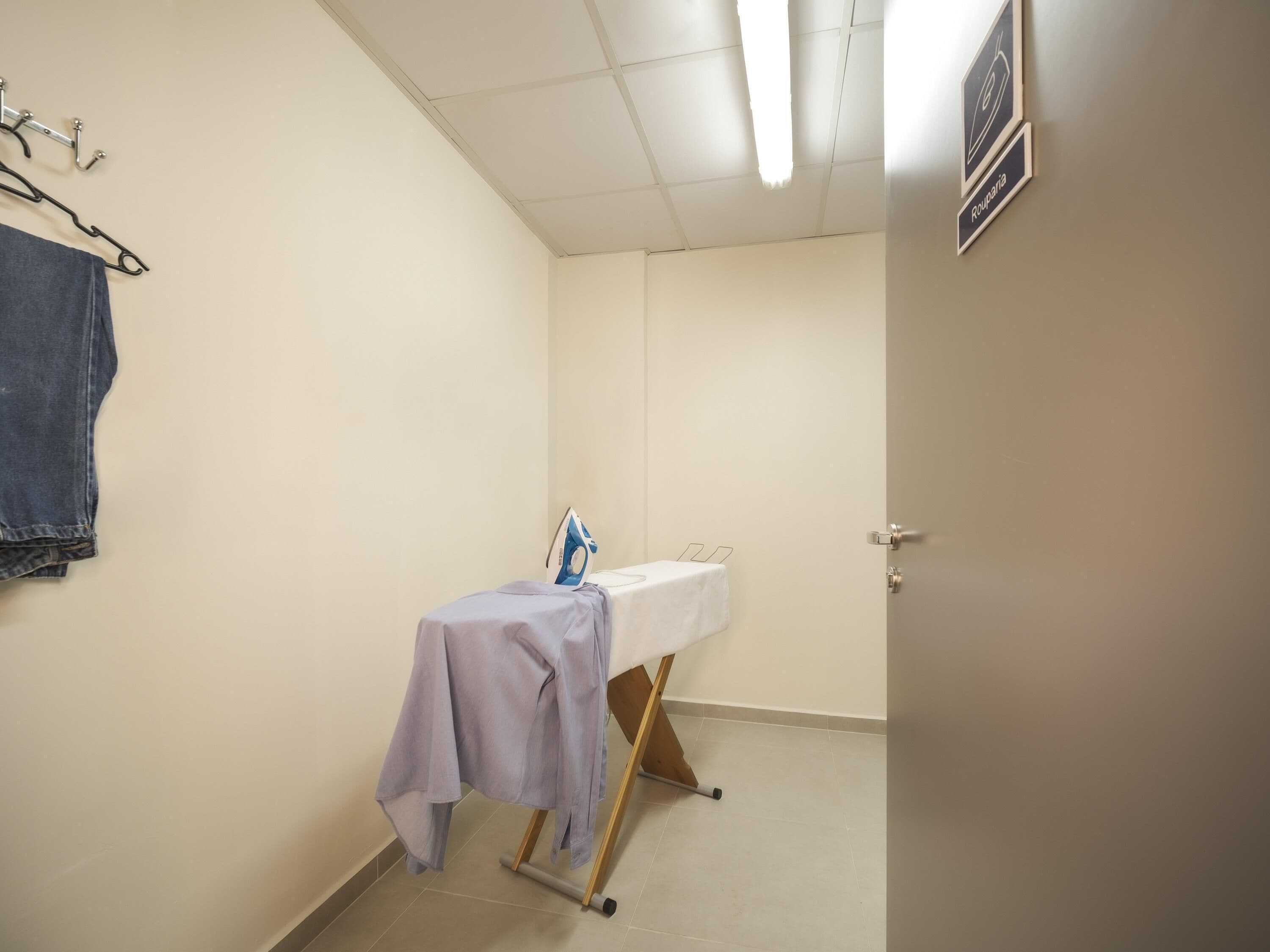 A small room with a bed, a table with an ironing board, a towel rack, and a door.