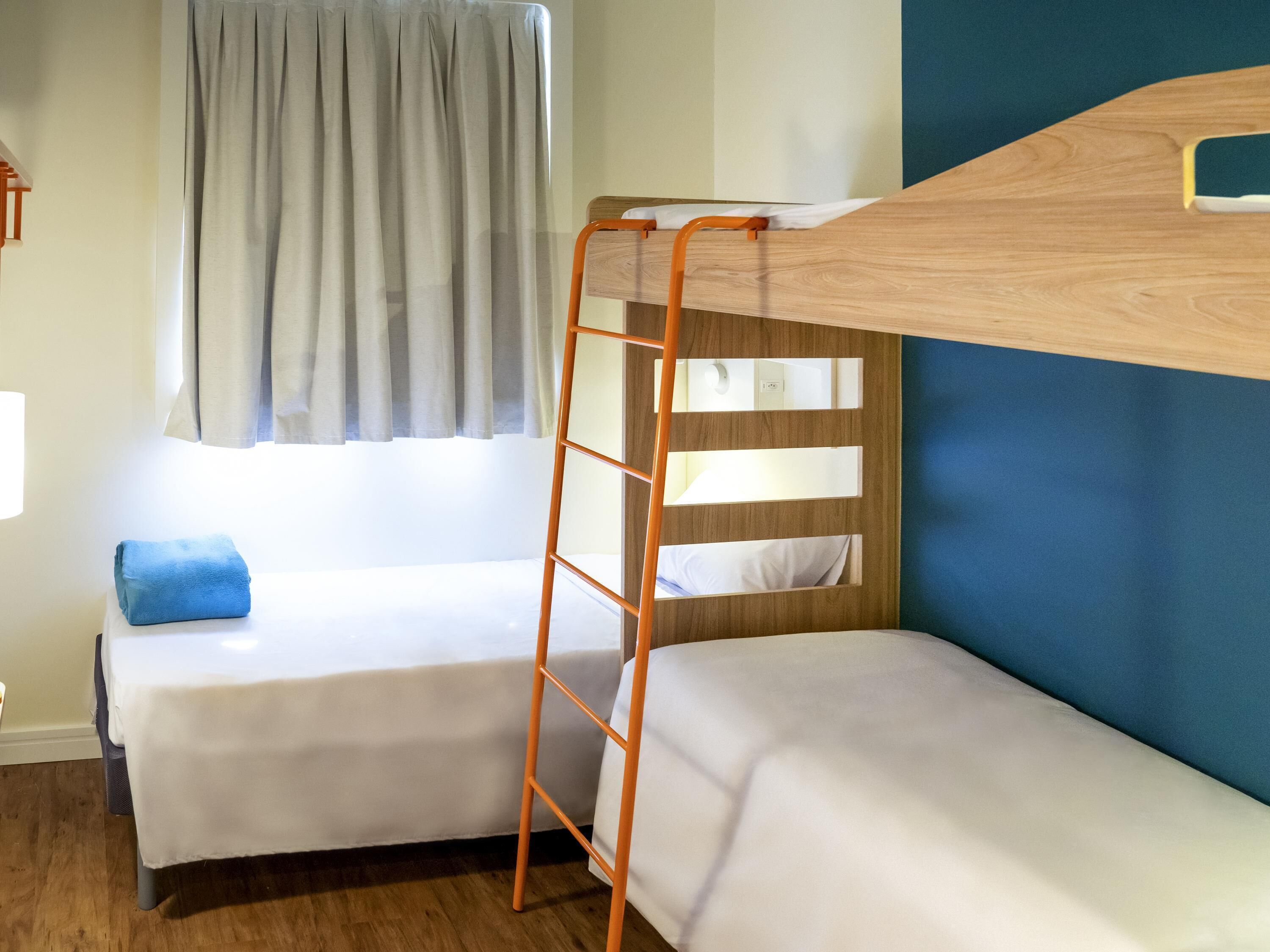 A bunk bed with a wooden ladder, a single bed with a pillow, and a window with curtains.