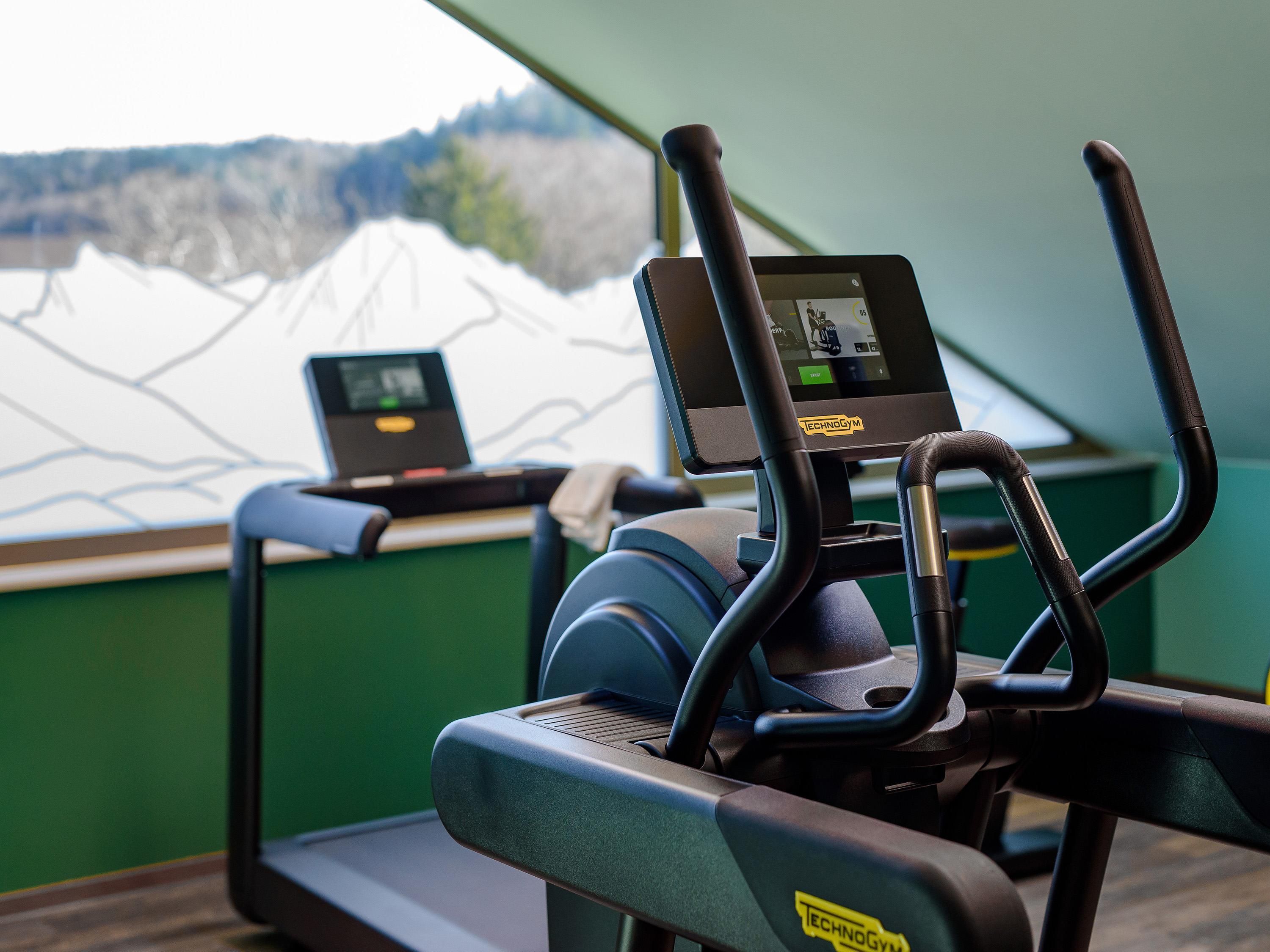 Un tapis de course doté d’un écran numérique, un ordinateur portable posé sur une table à proximité, et une vue sur des montagnes enneigées par la fenêtre.