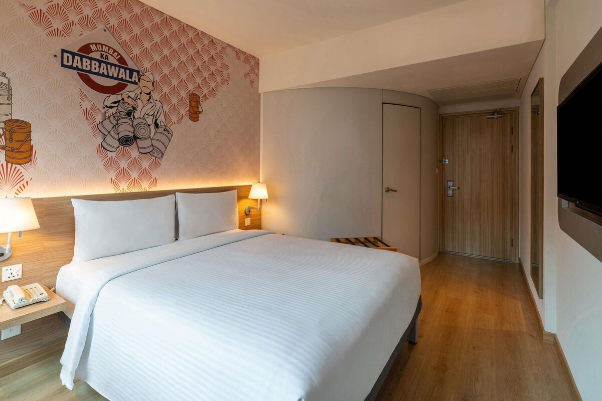 A hotel room with a bed, bedside lamps, a wardrobe, a television, and a wall with a Mumbai Dabawala logo.