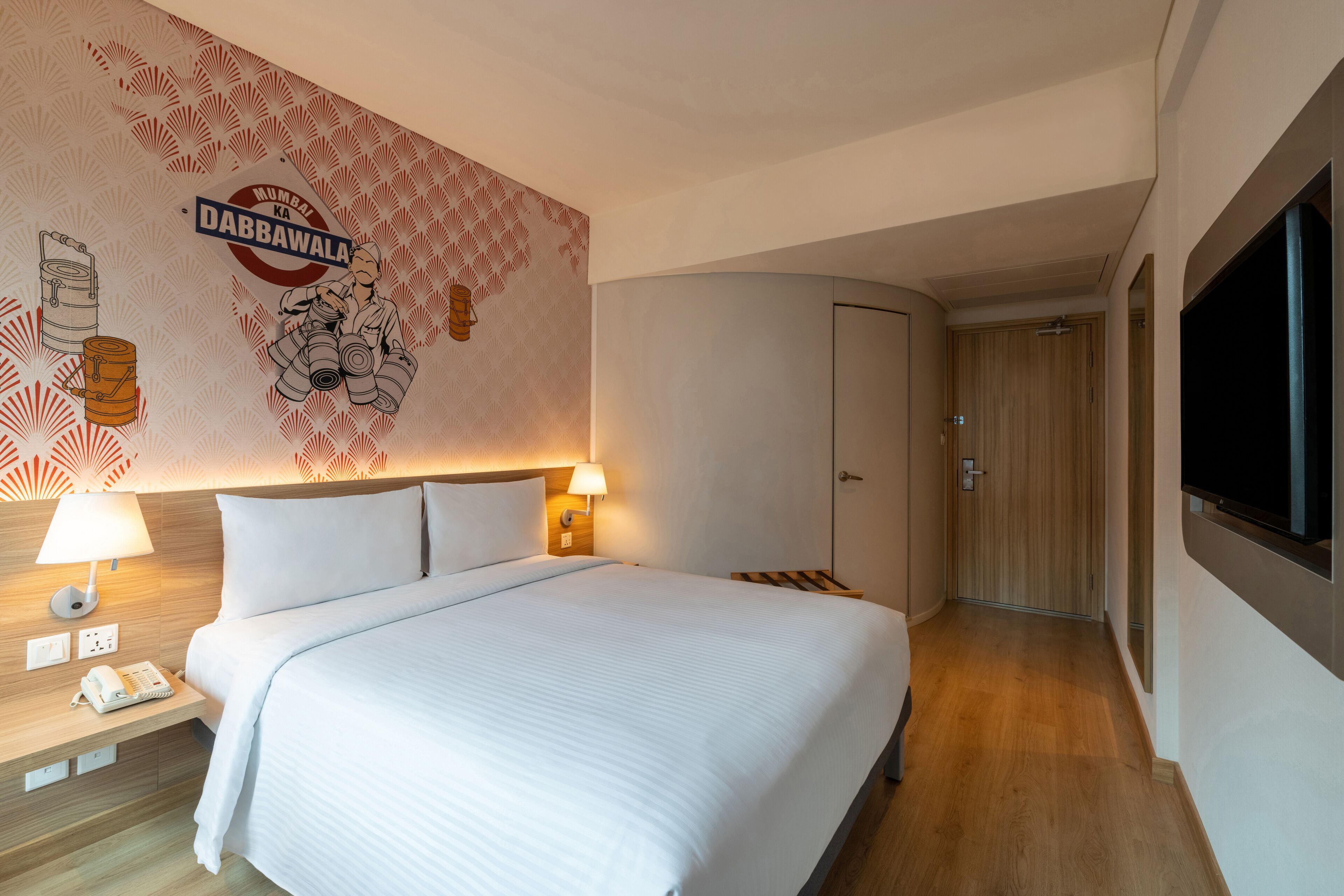 A hotel room with a bed, a nightstand with a lamp, a telephone, a wardrobe, and a television on the wall.