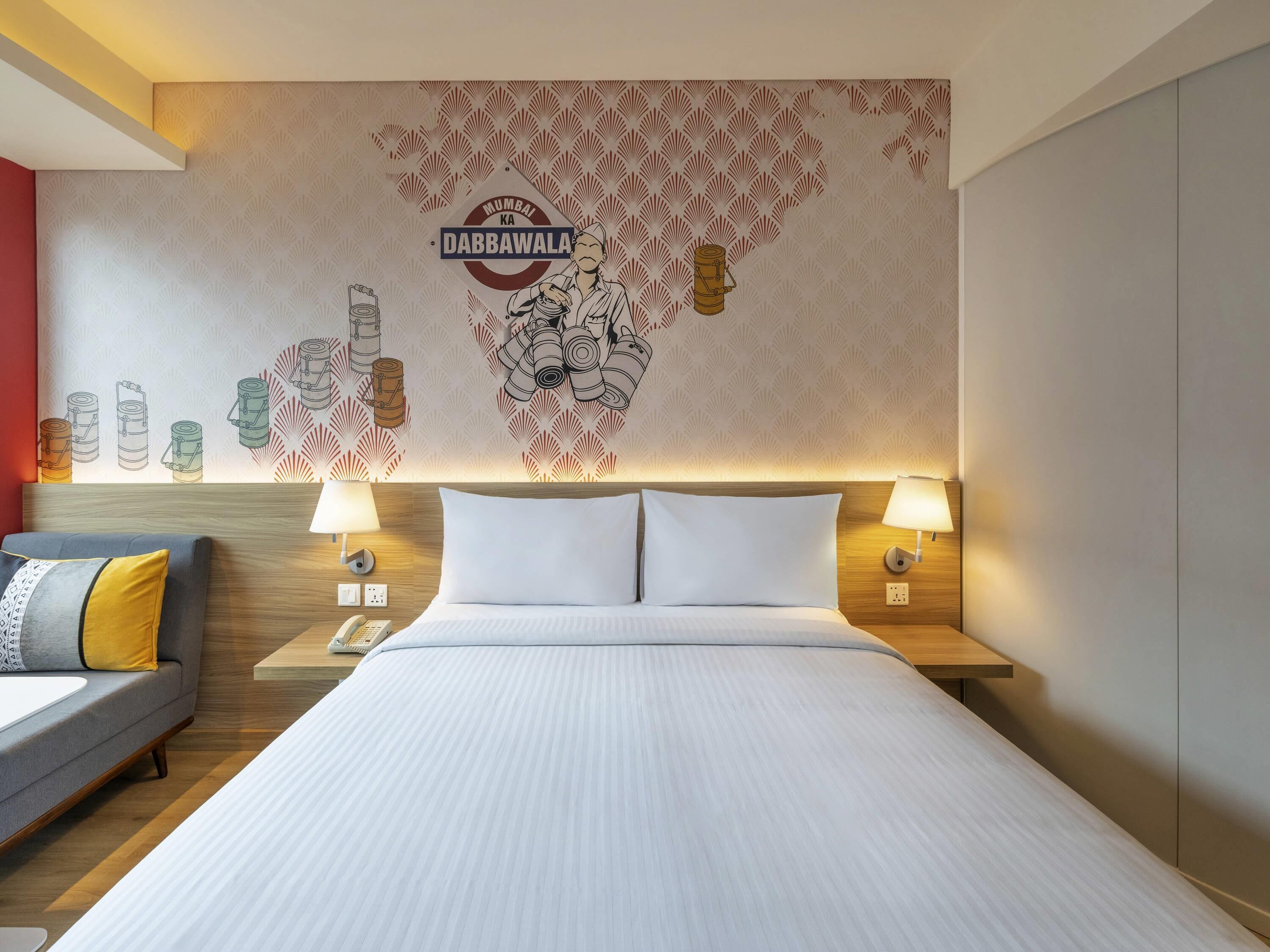 A hotel room with a bed, two bedside lamps, a nightstand, a sofa, and a wall with a Mumbai Dabbawala logo and illustrations of food items.