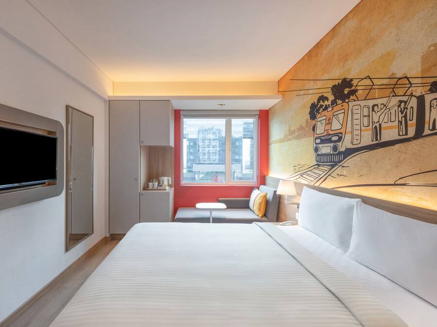 Een moderne hotelkamer met een groot bed, een aan de muur bevestigde televisie, een klein tafeltje en een bank met uitzicht op de stad.