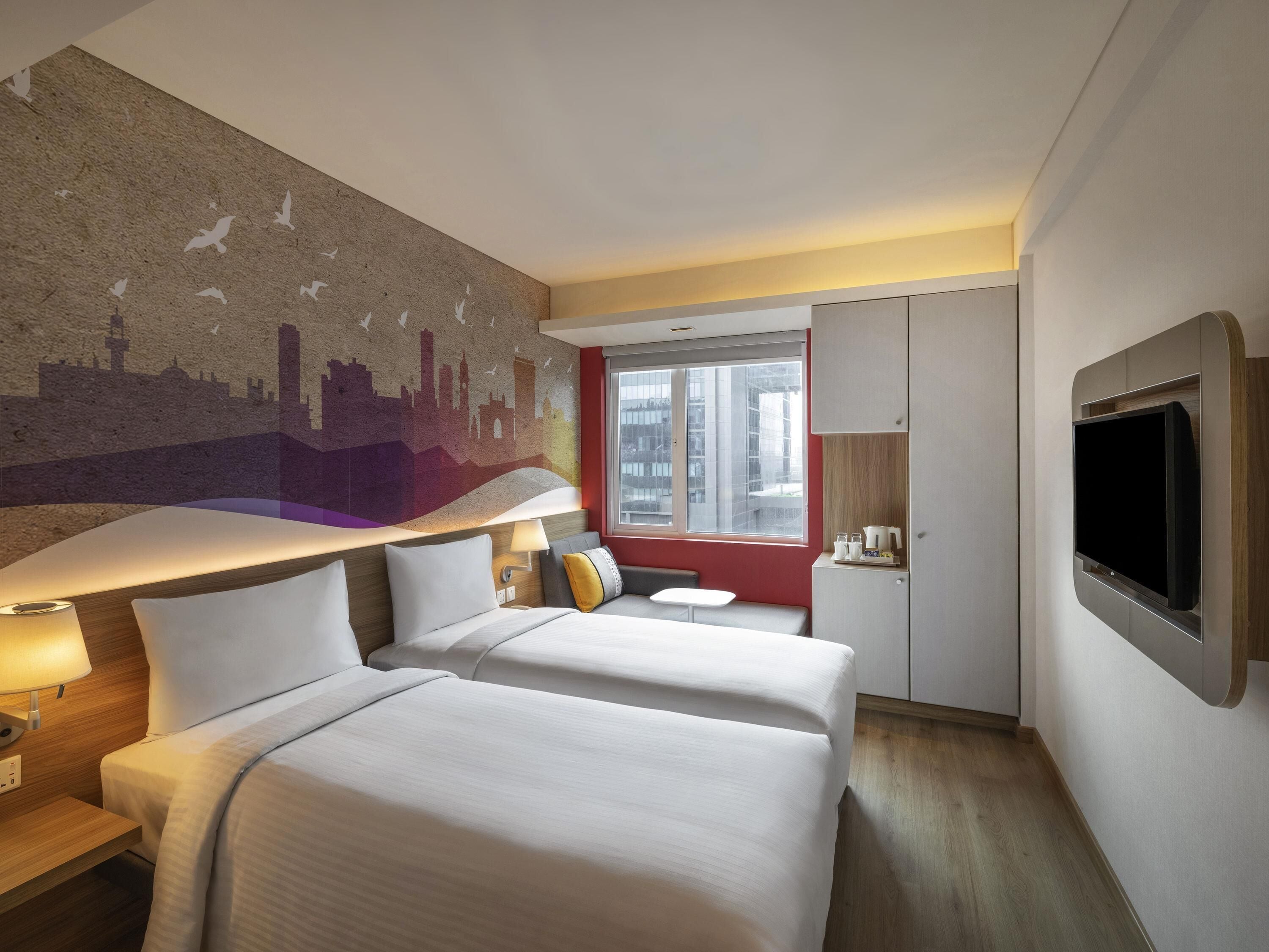A hotel room with two beds, a cityscape mural, a TV mounted on the wall, and a window with a view of buildings.