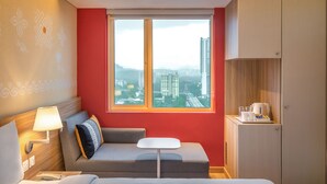 Une chambre d’hôtel avec un canapé, une petite table, une lampe, une fenêtre donnant sur la ville et un meuble sur lequel sont posés divers objets.