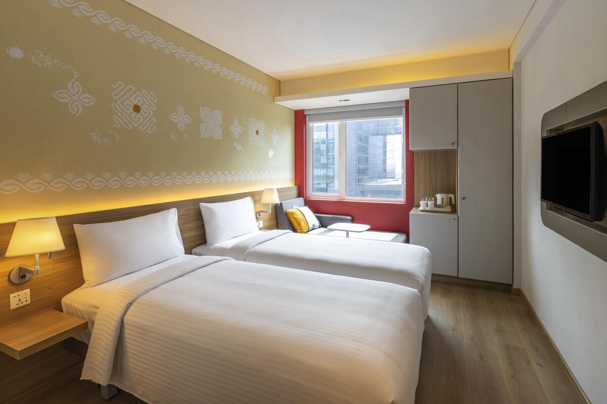 A hotel room with two beds, a window with a city view, a TV mounted on the wall, and a small table with a lamp.