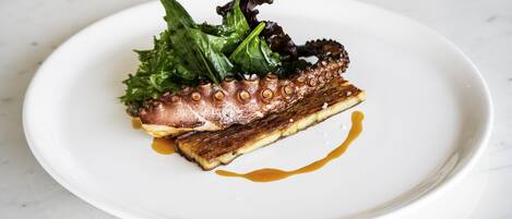 A white plate with octopus, greens, and a sauce on a marble surface.