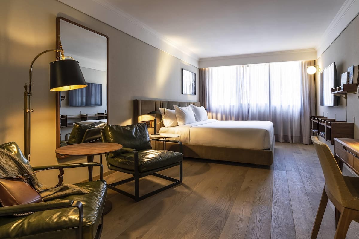 Una habitación de hotel con cama, escritorio, silla, lámpara y un espejo grande.
