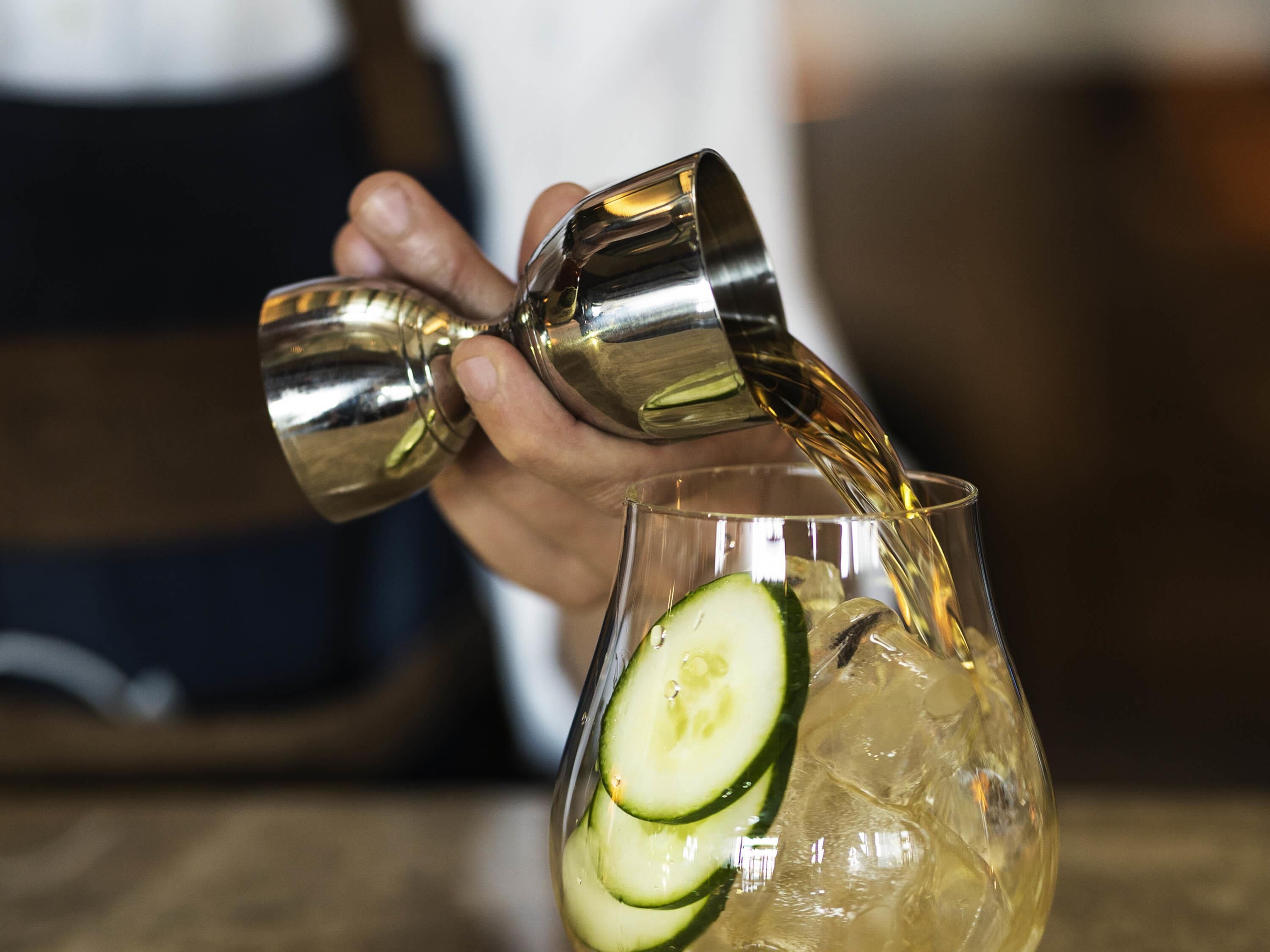 A person pouring a golden liquid into a glass with ice and cucumber slices.