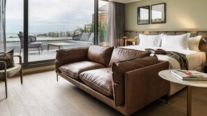 Ett modernt hotellrum med ett stort fönster, en lädersoffa, en säng med vita sängkläder och ett litet runt bord med en bok på.