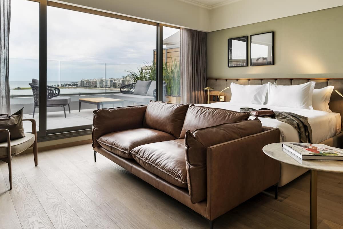 Una habitación de hotel moderna con un ventanal grande, un sofá de cuero, una cama con sábanas blancas y una pequeña mesa redonda con un libro.