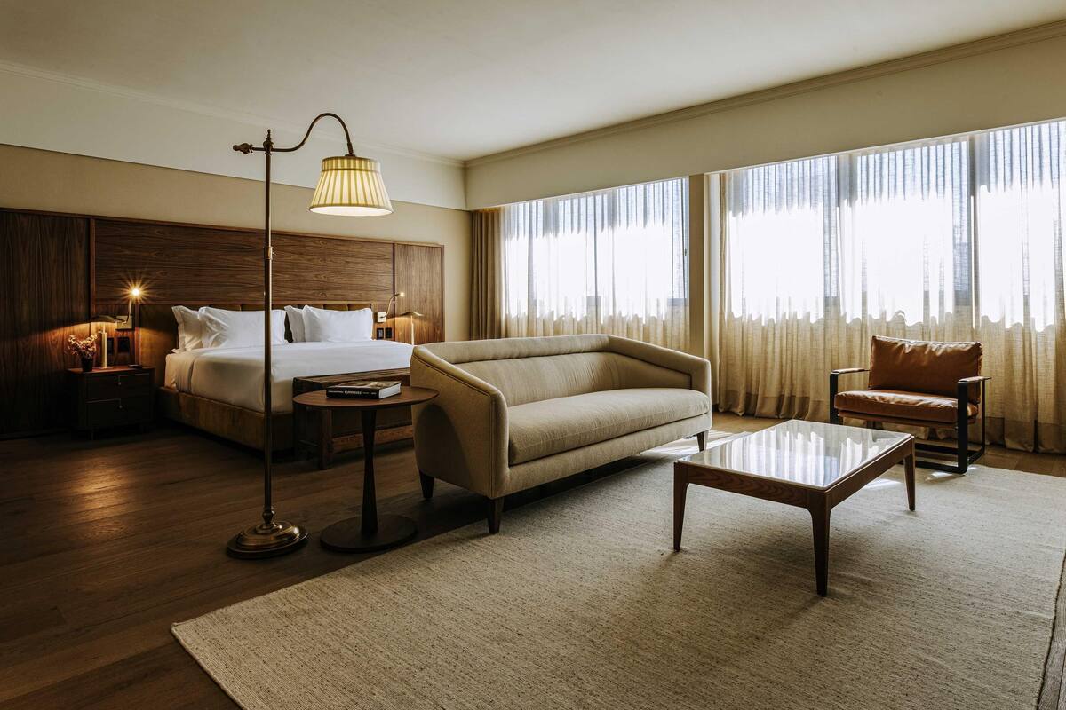 Habitación de hotel con cama, sofá, sillón, lámpara de pie, mesita de noche y ventanas amplias.