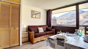Ein gemütliches Zimmer mit einem Holzschrank, einem braunen Sofa, einem kleinen Tisch mit weißer Tischdecke und einem Fenster mit Blick auf die Berge.