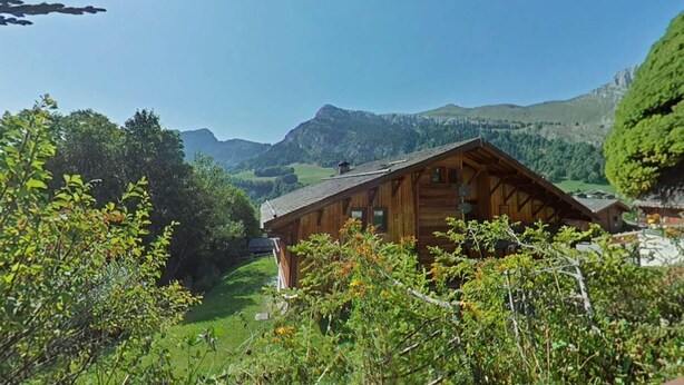 Eine Holzhütte mit geneigtem Dach, umgeben von üppiger Vegetation und Bergen im Hintergrund.