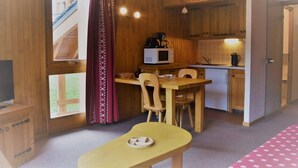 Habitació senzilla amb interior de fusta, una cuineta petita, zona de menjador amb taula i cadires, i finestra amb cortines.