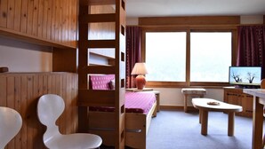 Habitació amb lliteres, escriptori, cadira, televisió i finestra amb cortines.