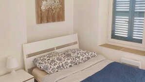 Ein Schlafzimmer mit Bett, Nachttisch, Fenster mit Jalousien und einem Wandbild.