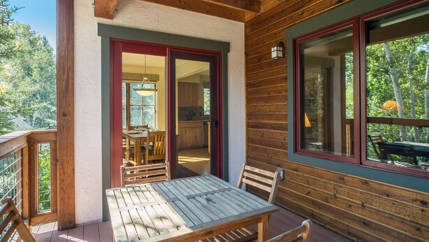 A wooden deck with a table and chairs, a sliding glass door leading to a kitchen, and a view of trees. - The Pines 202 2 Bedroom Condo by RedAwning (Steamboat Springs)