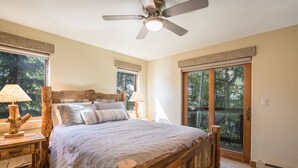 A bedroom with a wooden bed, two bedside lamps, a ceiling fan, and large windows with a view of trees. - The Pines 202 2 Bedroom Condo by RedAwning (Steamboat Springs)
