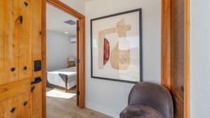A hotel room with a wooden door, a framed abstract artwork on the wall, and a bed with a white cover. - @ Marbella Lane - Rainbow House | Glampers Paradise (Joshua Tree)
