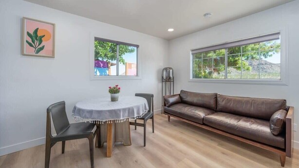 A modern living room with a brown leather sofa, a round dining table with two chairs, and large windows offering views of greenery and buildings. - @ Marbella Lane - Rainbow House | Glampers Paradise (Joshua Tree)